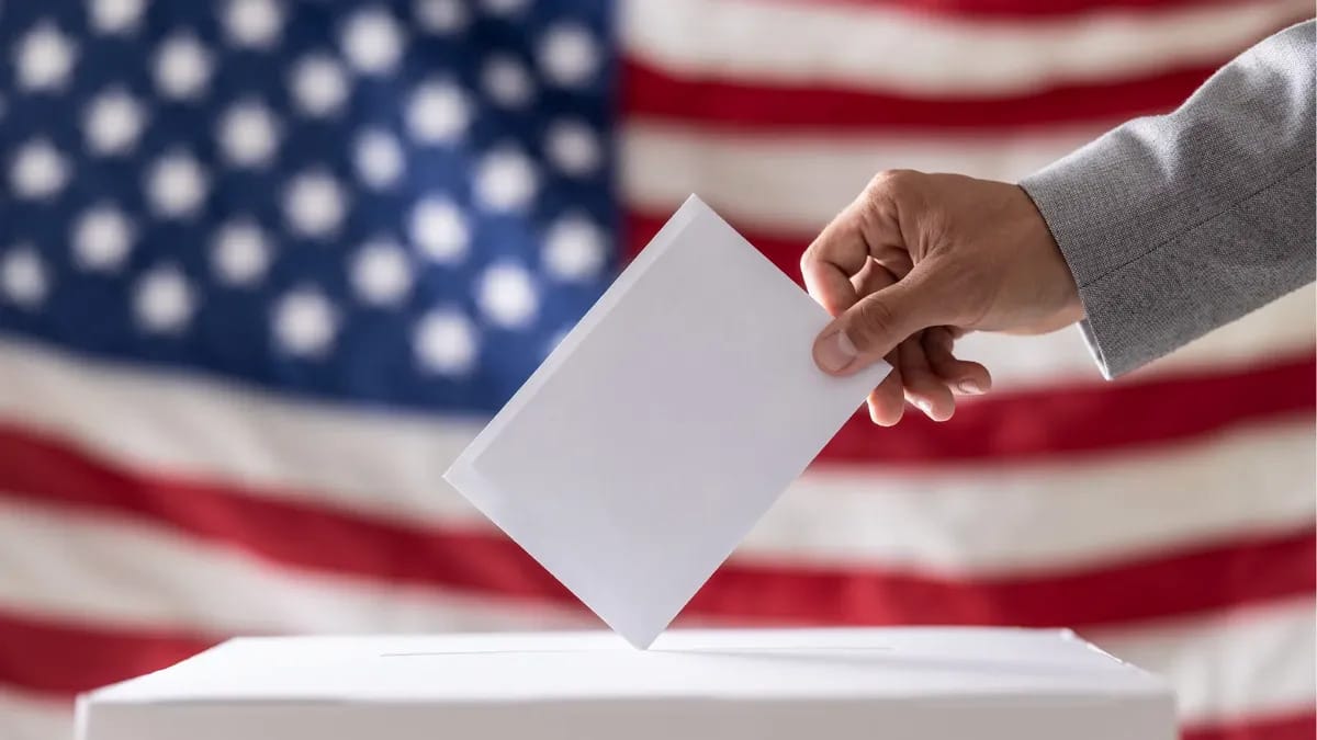 A person places an envelope into what appears to be a ballot box. A red, white and blue flag can be seen in the background.