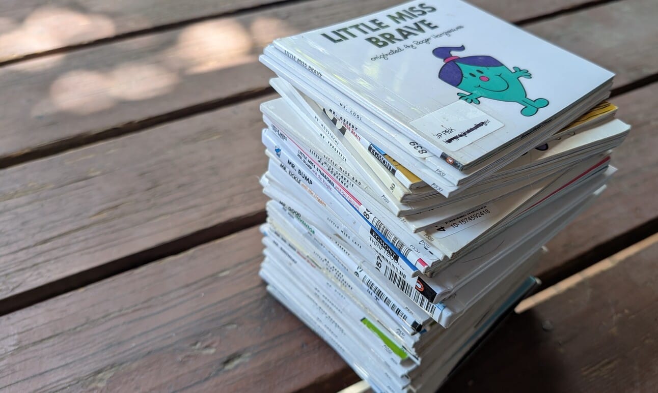 A stack of about 50 square white books sit on a wooden deck. The top book says "Little Miss Brave" and features a small teal cartoon person with purple hair. 