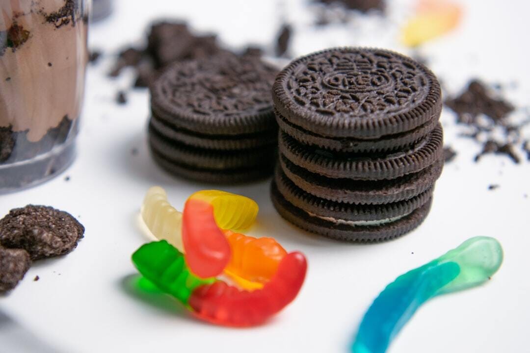 A close-up shot of a hand placing a gummy worm on top of a dirt pudding cup layered with chocolate pudding and crushed cookies. The dessert is served in a clear plastic cup, emphasizing texture and color. This playful food image is well suited for kids’ recipes, party desserts, Halloween treats, or nostalgic food content.