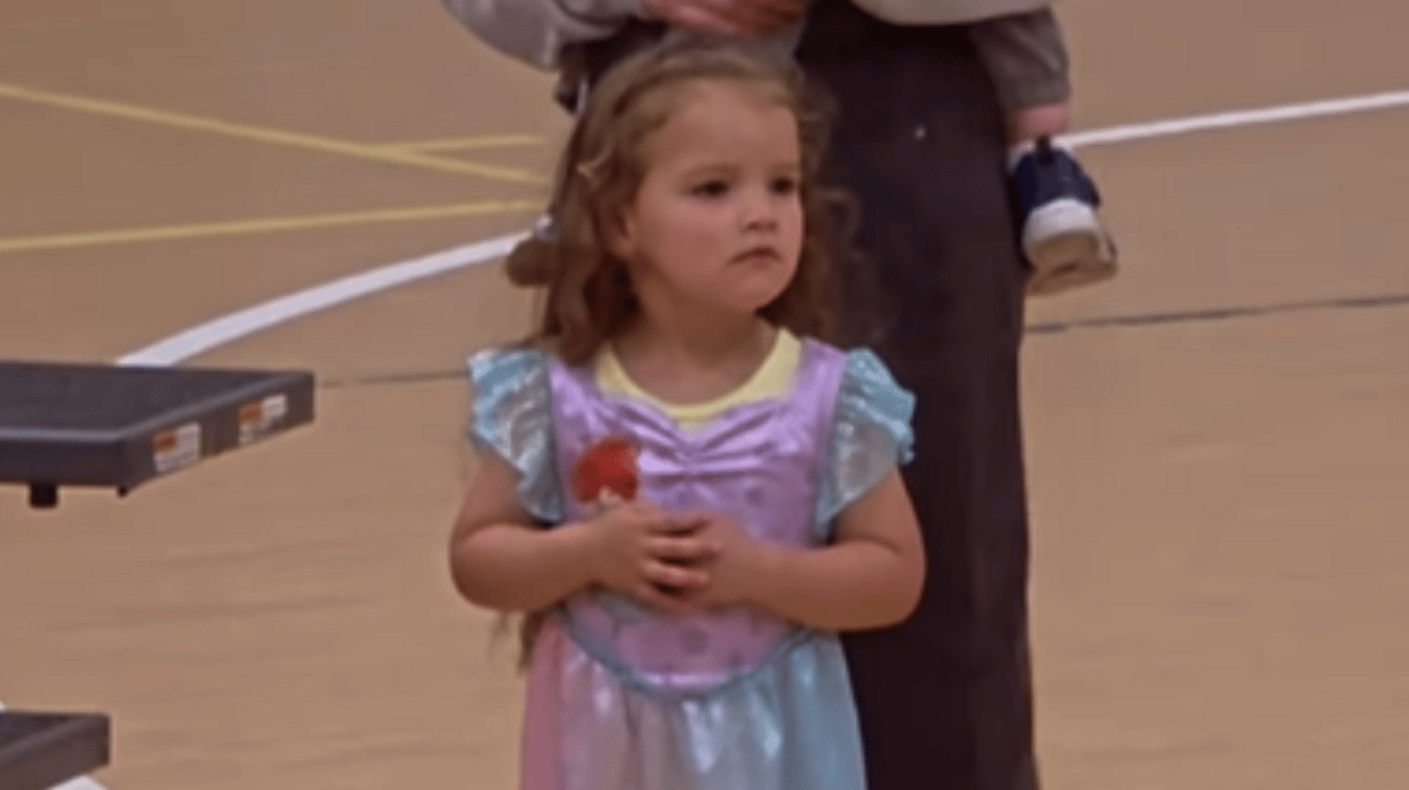 A little girl stands by an adult, looking around nervously. There's a small frown on her face as she holds her own hands in front of her stomach