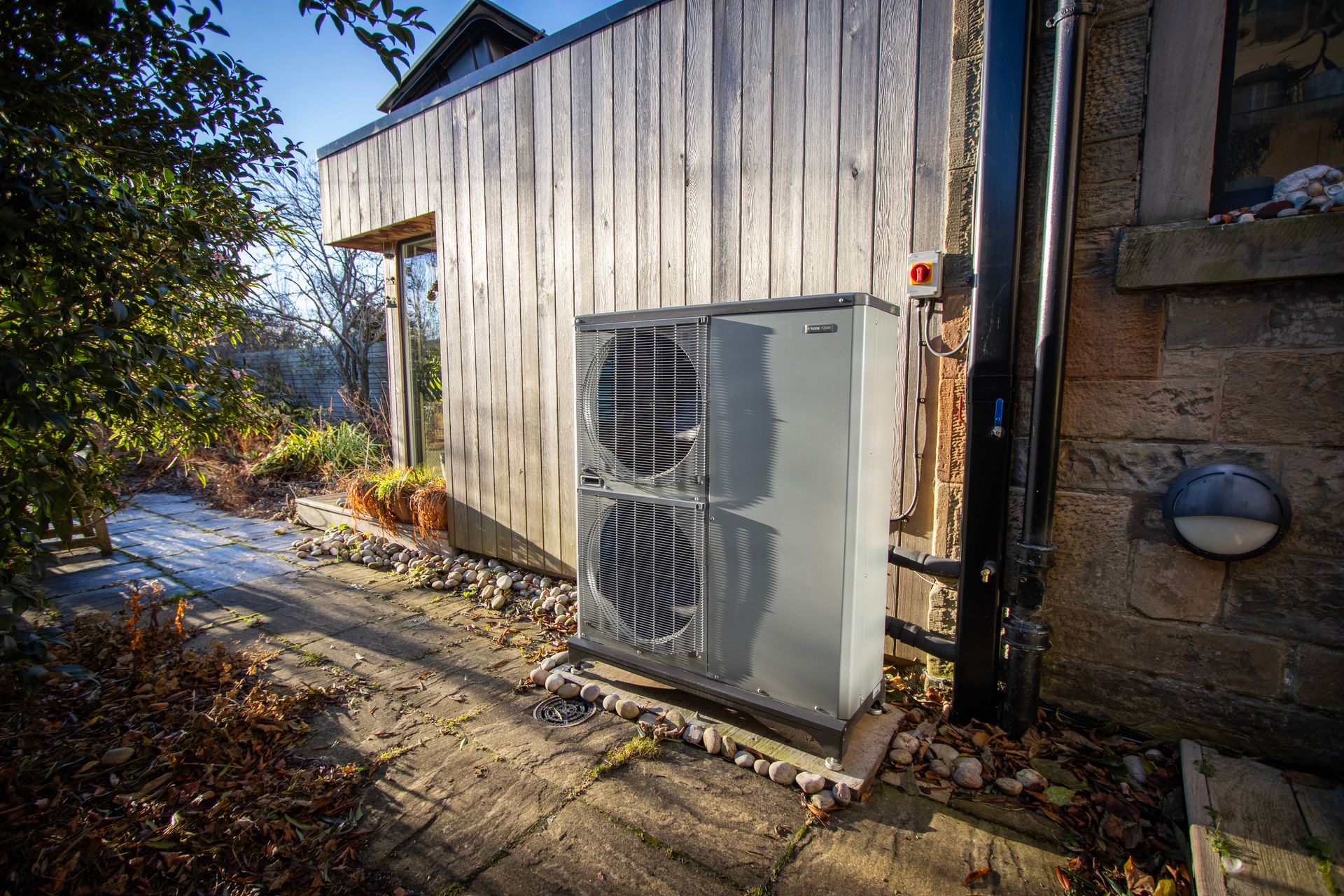 A Nibe F2040 16kW air source heat pump installed outside a traditional pre-1919 sandstone semi-detached home in Edinburgh. The unit is mounted on a stable base with anti-vibration feet next to a timber-clad extension and stone wall, featuring professional pipework and electrical connections.