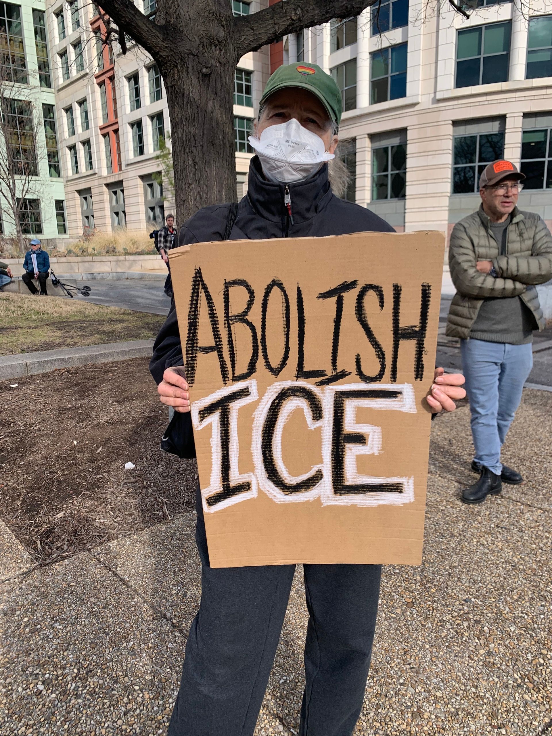 A woman with her hair pulled back is wearing a baseball cap with a rainbow heart on it and holding a cardboard sign that reads "ABOLISH ICE." She is standing on a city street with a tree visible in the background.