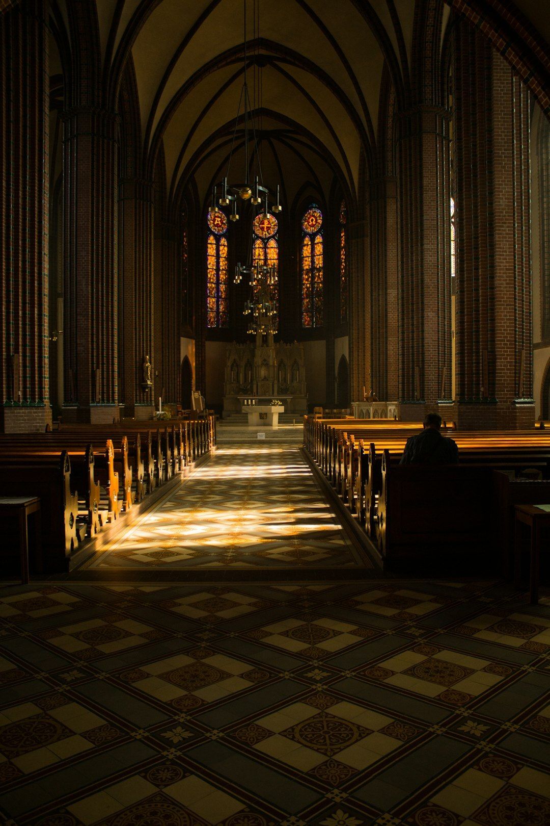 In a church with high arched ceilings, stained glass windows and long wooden pews, a man sits in one of the pews in the back, bowing his head.
