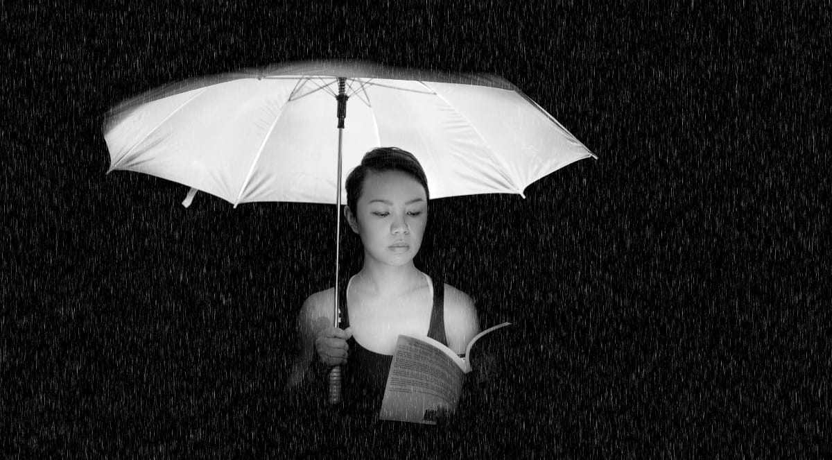 foto em preto e branco de uma mulher lendo um livro debaixo de chuva enquanto segura um guarda-chuva branco. Ela é uma mulher de pele clara com traços do leste asiático, está com o cabelo preso e veste uma regata preta.
