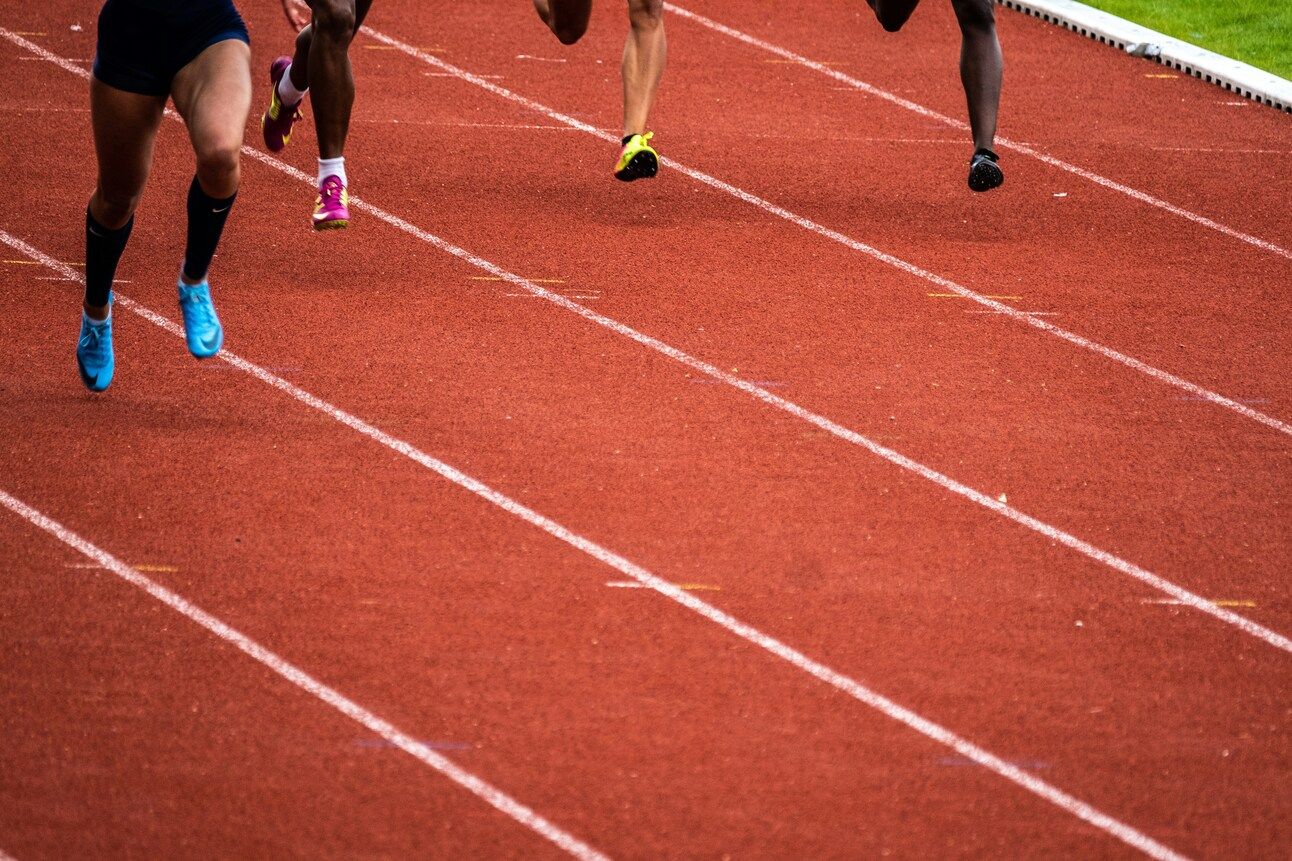 The legs of four different runners are seen on a clay-coloured running track