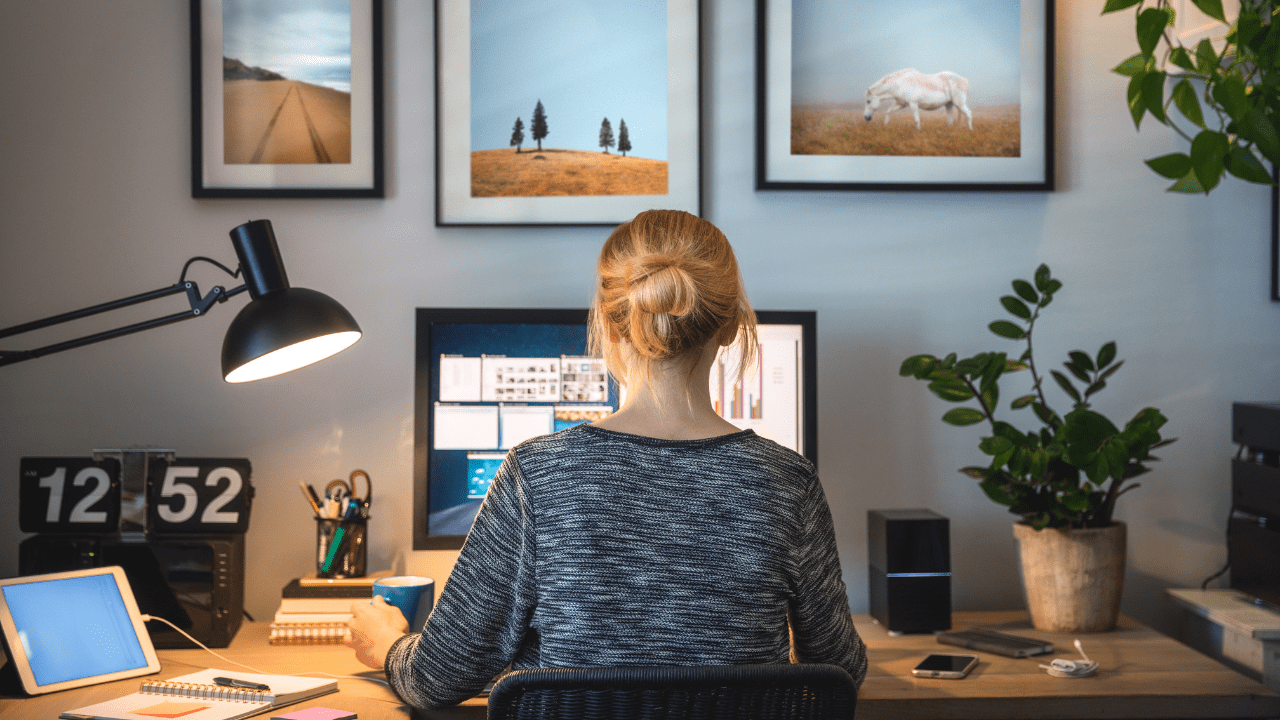 Woman sitting at a neatly organized desk working on a computer with framed photos of nature scenes on the wall behind her.