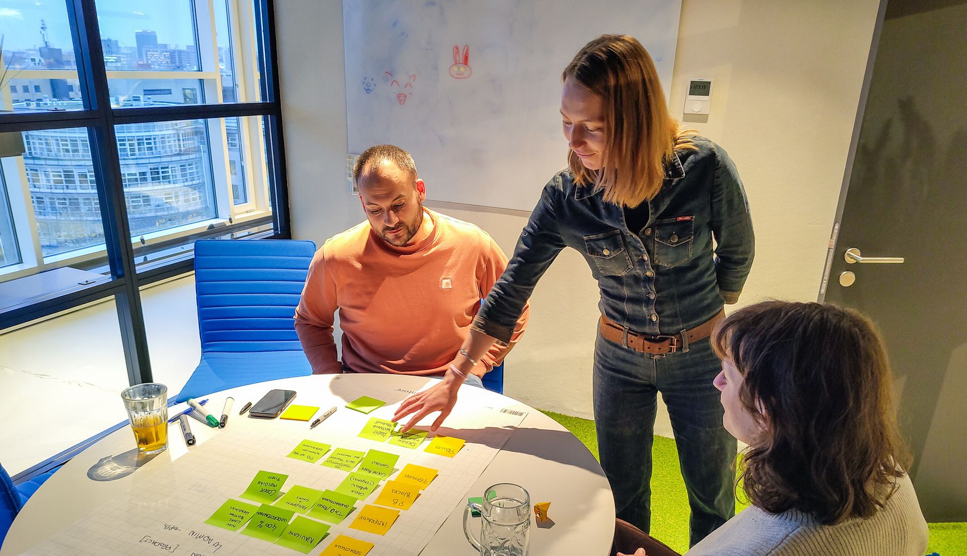 Twee mensen zitten aan een ronde tafel met post-its, Emma van Digitaal Toegankelijk staat bij de tafel en gebaart naar de post-its