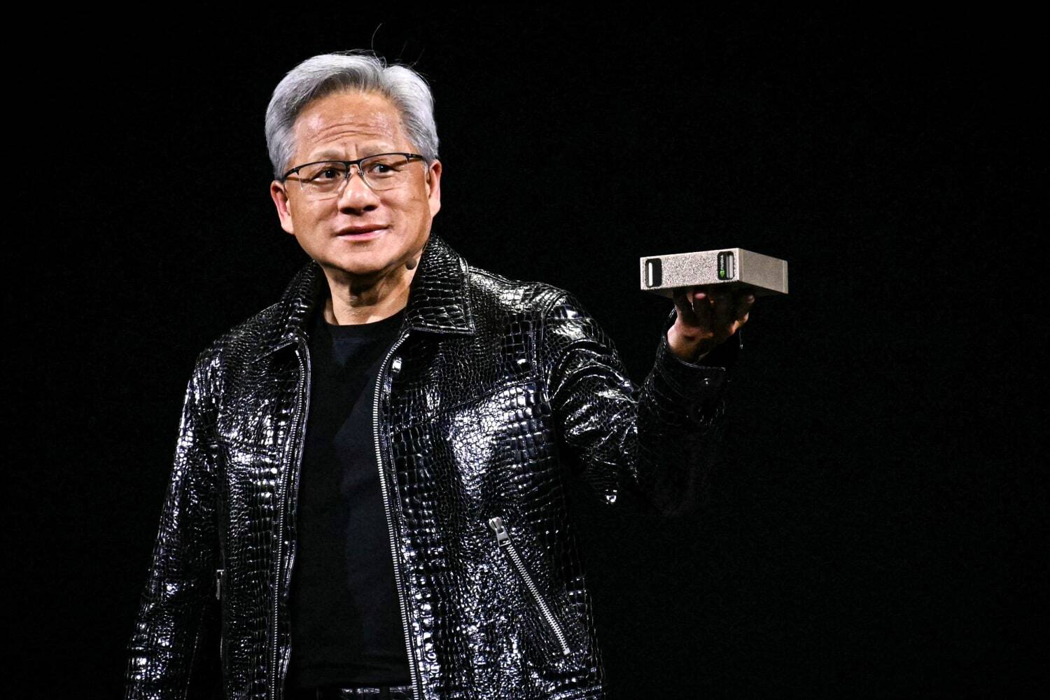 An in-depth, eye-level, medium shot shows Nvidia CEO Jensen Huang on a stage with a solid black background. He is a man with short, graying hair and wears glasses. He is turned slightly to his left while looking towards the right side of the frame with a focused, confident expression. He is wearing his signature black leather jacket, which has a glossy, textured, snakeskin-like pattern. In his left hand, he is holding up and presenting a small, rectangular, metallic silver device, likely a piece of computer hardware. The stage lighting illuminates him from the front, highlighting the details of his face, his jacket, and the device he is holding.