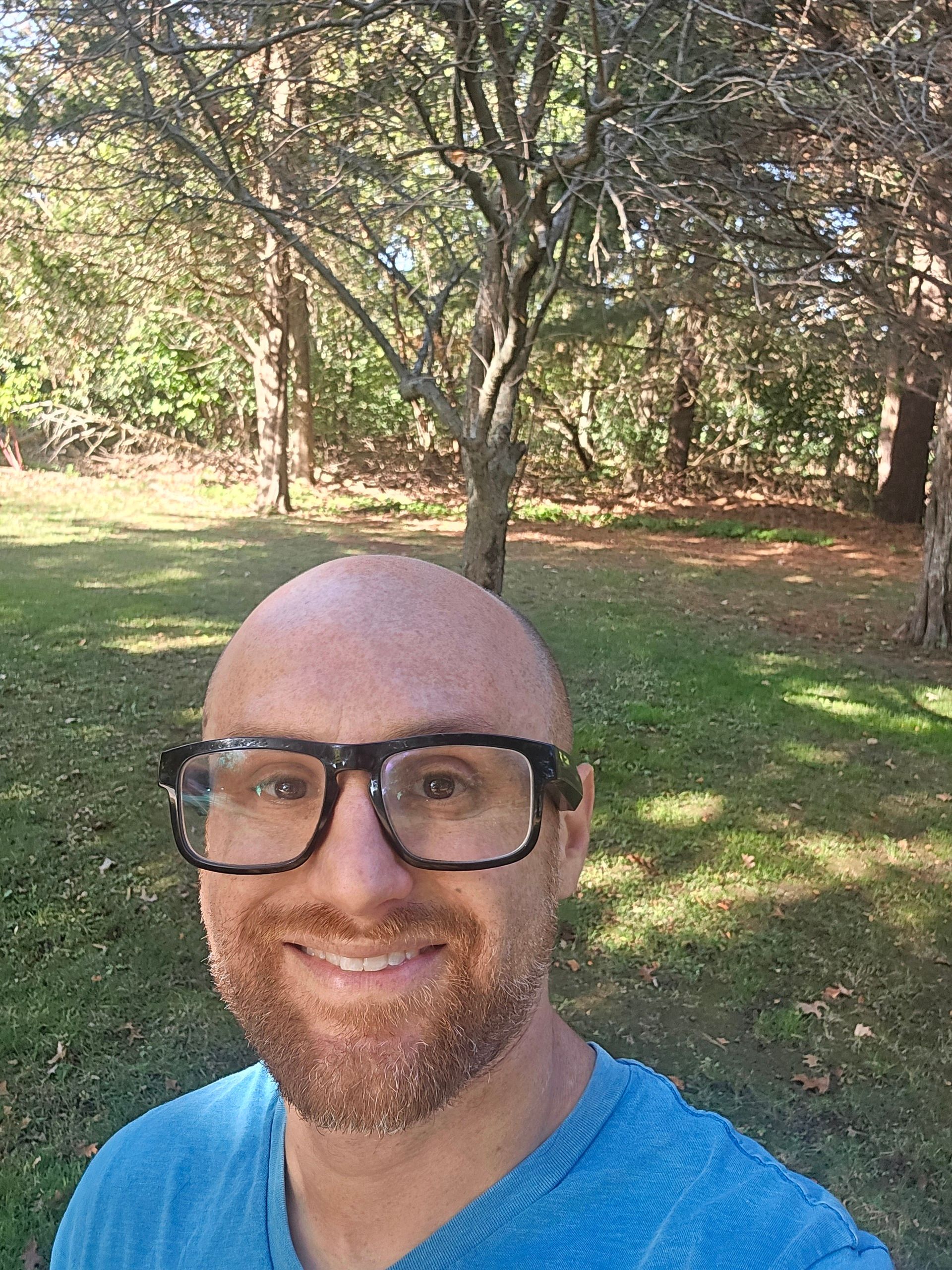 A selfie of me on a grassy lawn in front of forest on a sunny day. I'm a bald, middle-aged, white man with a red beard flecked with white. I'm wearing glasses with thick, black rims and a bright blue shirt.