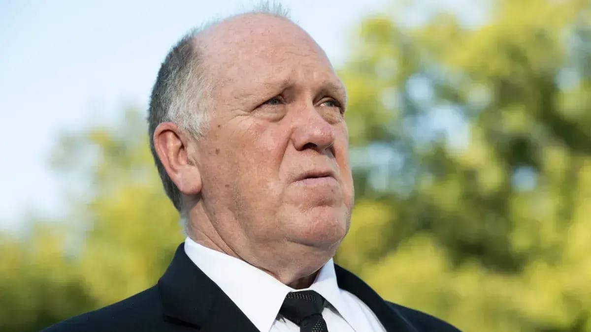 A white man wearing a black suit looks towards the right.