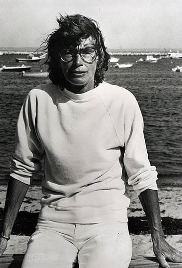 Mary Oliver sitting on a wooden railing in front of Provincetown Harbor, her arms muscled, her hair in her face.