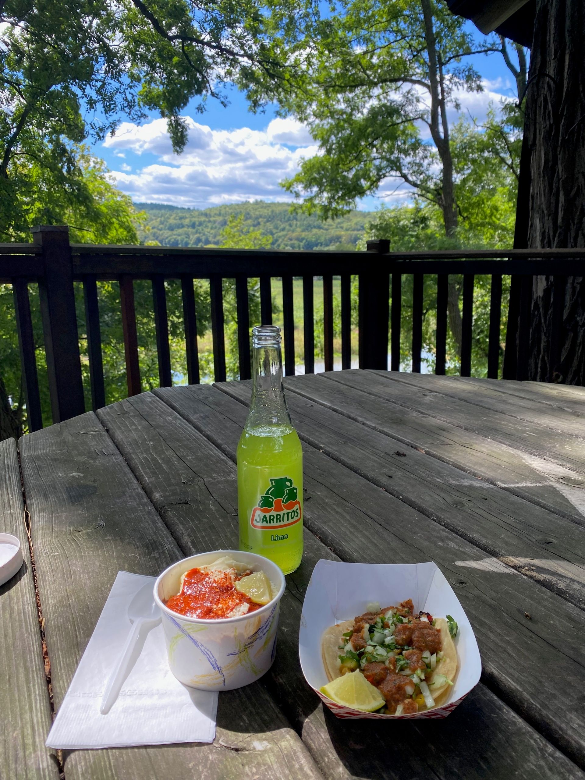 Tacos, elote, and a lime soda on a picnic table overlooking the river, wetlands, hills, and trees.