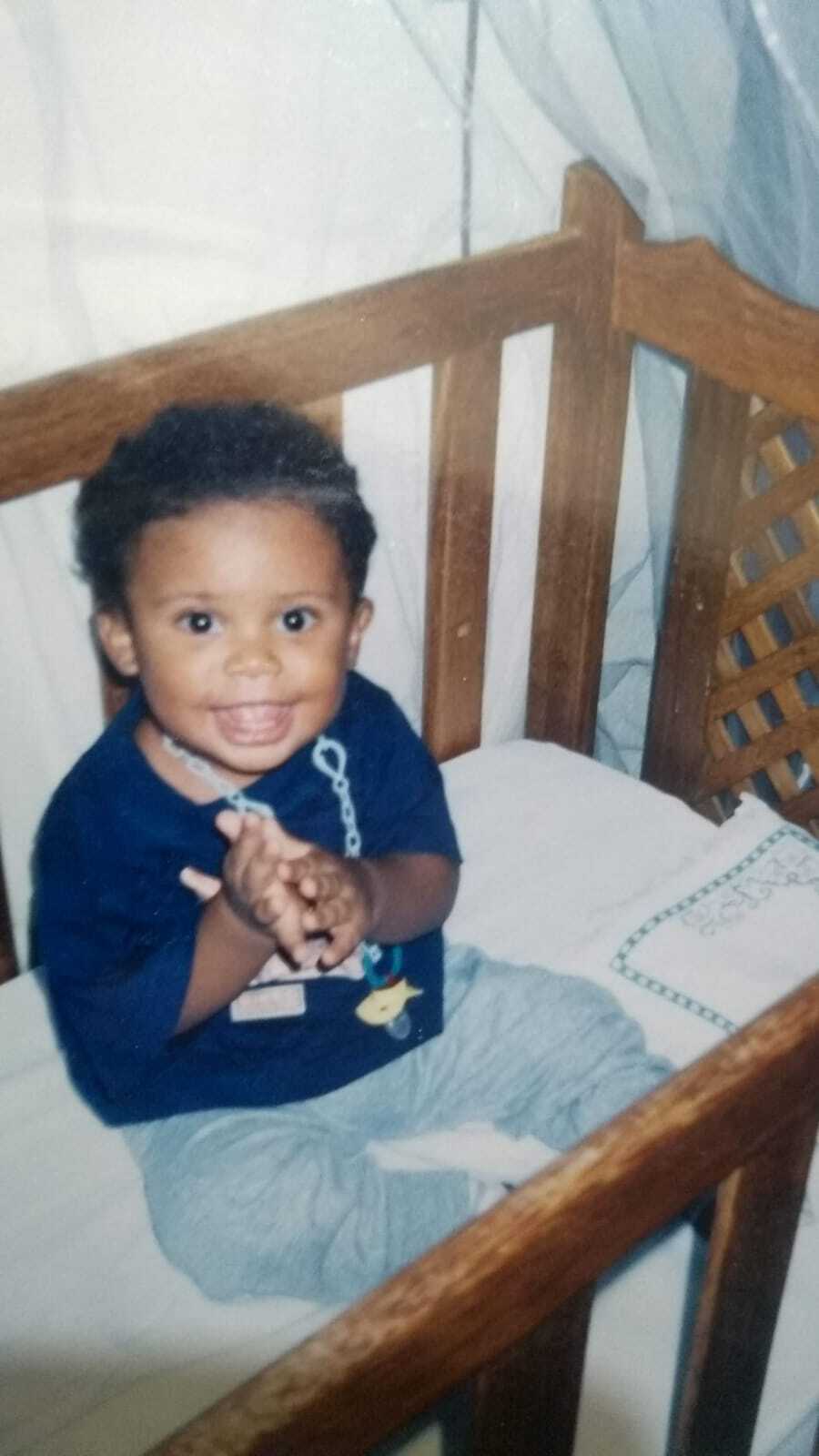 Um bebê negro no berço sorrindo e com as mãos juntas.