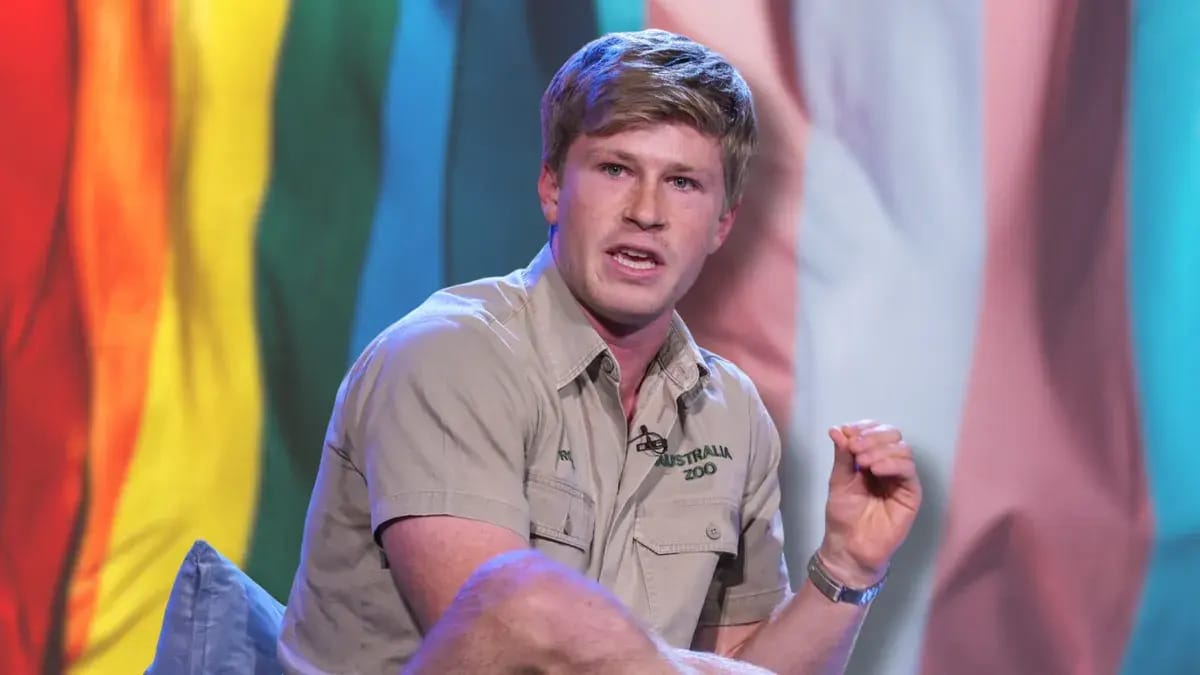 A white man is sitting down wearing a shirt that says AUSTRALIA ZOO. In the background, a pride flag is shown, which has colors of the rainbow on it next to pink, white and blue stripes to the right.