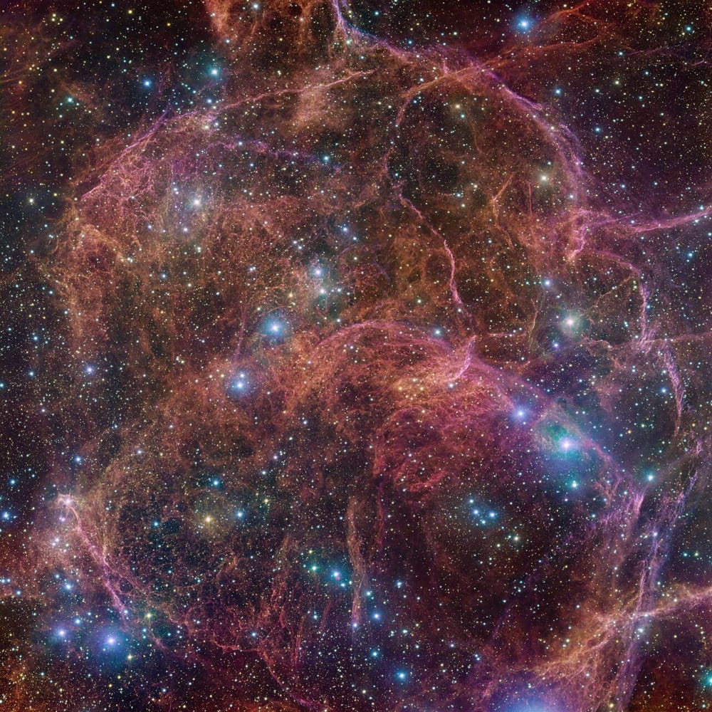 An image showing thousands of stars in a black sky, and among them are dozens, if not hundreds, of then filaments of red and pink gas distributed in a roughly spherical shape.