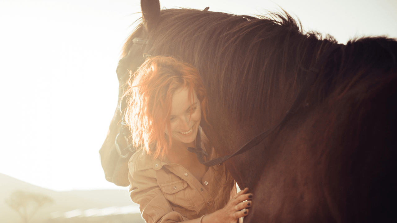 Smiling woman with red hair affectionately hugging a brown horse in warm golden sunlight.