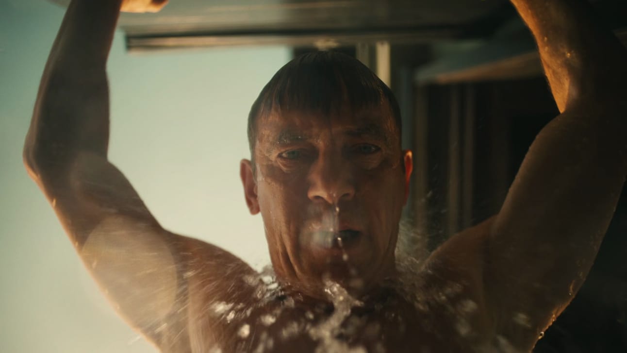 Jason Isaacs stands in a shower, arms baced over his head, water ouring down