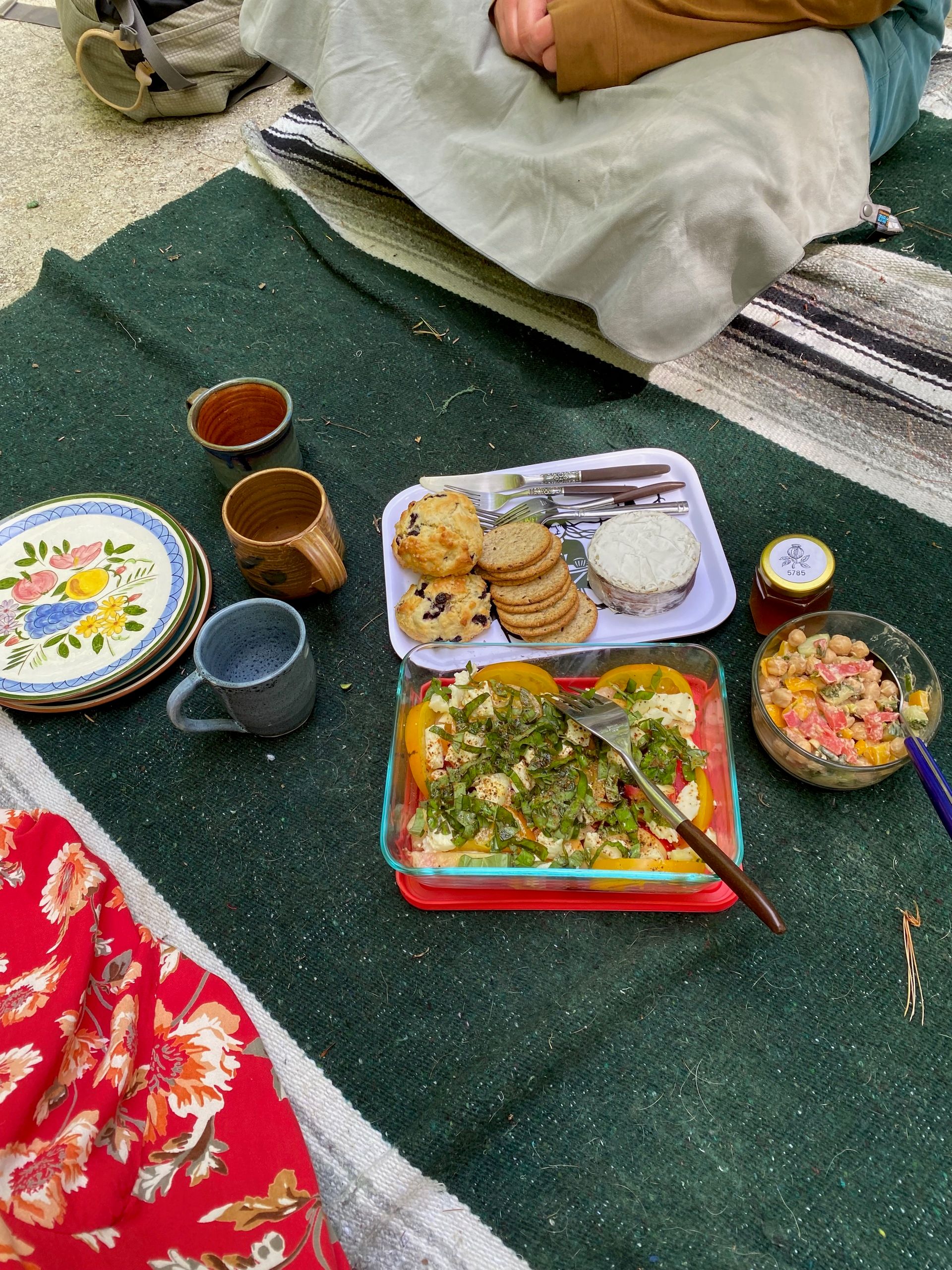 A spread of crackers, cheese, scones, a tomato-melon salad, and ceramic mugs and plates on a blanket on a rock.