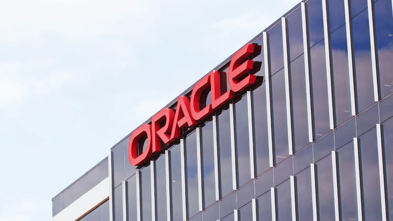 Photo of the Oracle headquarters with the company’s red logo mounted on a glass office facade reflecting the sky.