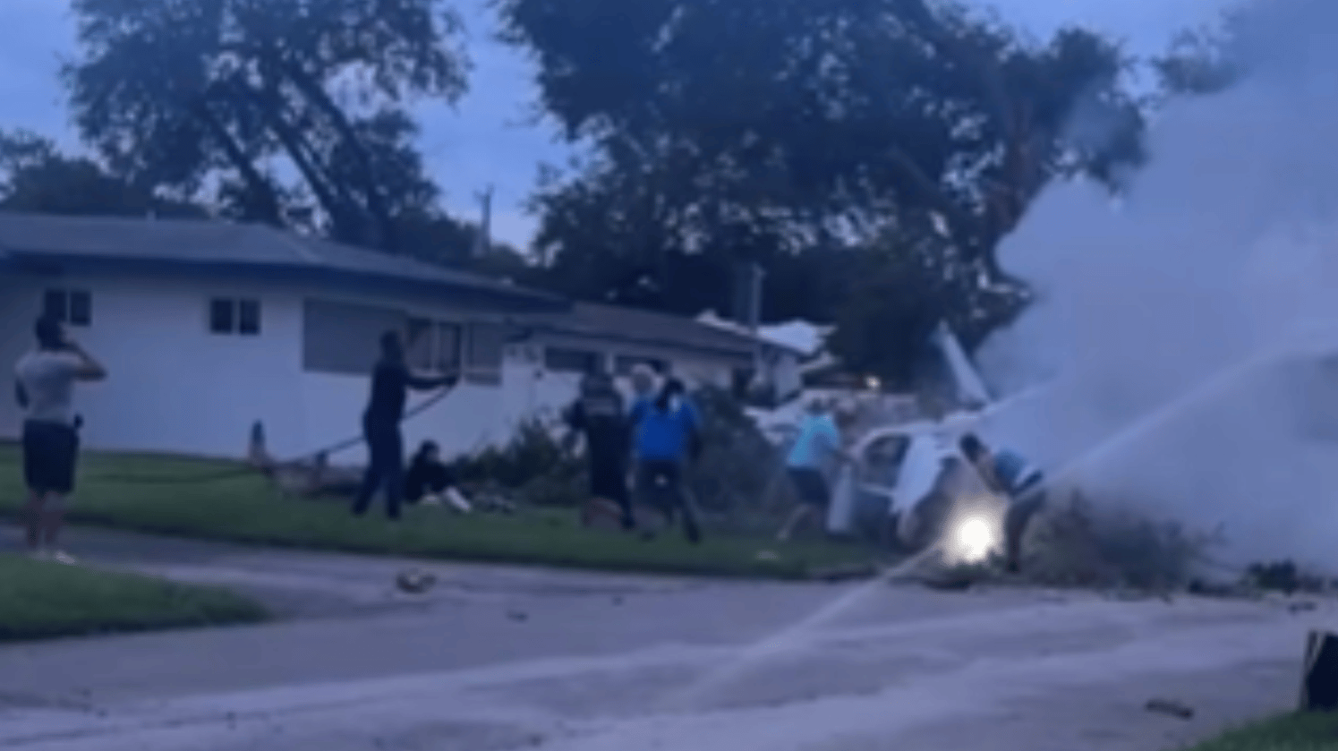 View from a distance of the aftermath of a plane crash. A whole group of people gather around to help rescue the people inside the plane. One person stands off to the side with a hose directed at the plane.