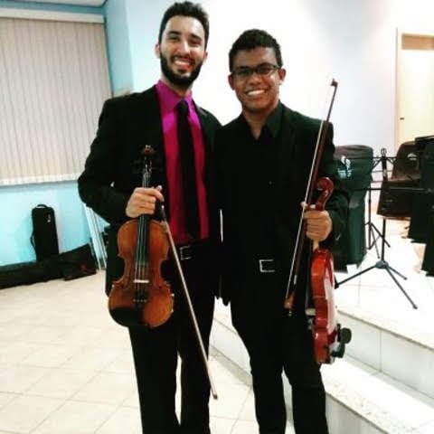 Dois homens segurando violinos. O da esquerda é mais alto, está com um terno e camisa rosa com gravata, tem pele branca e tem barba e cabelos pretos lisos. O da direita sou eu, om terno preto e camsa preta sem gravata, pele marrom escura, óculos, e cabelos crespos curtos.
