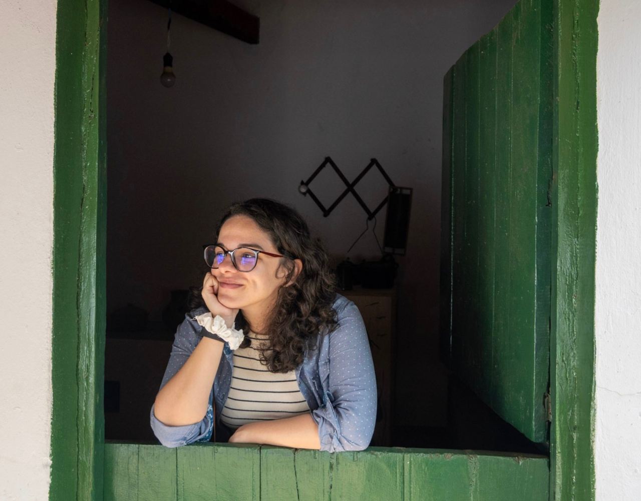 Fotografia da autora Fernanda Castro, uma mulher branca de cabelos cacheados pretos. A autora está apoiada numa porta verde, com o queixo apoado numa mão enquanto sorri para um ponto longe da câmera.