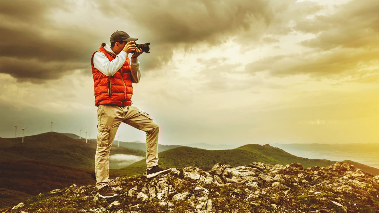 Photographer standing on a rocky mountain peak at sunrise, wearing a red vest and taking a picture of the landscape below.