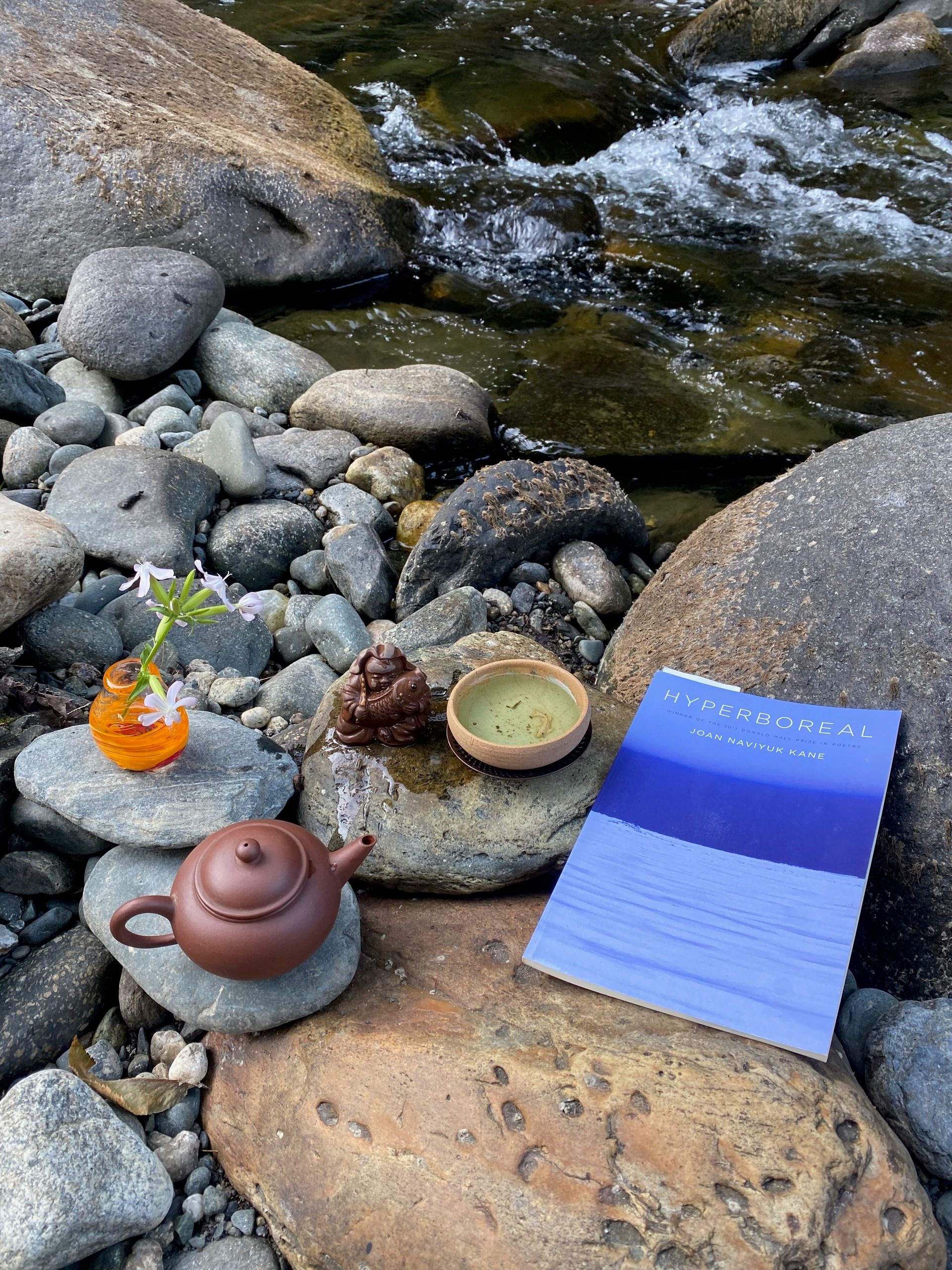 A small tea pot, cup, tea friend, tiny vase with a flower, and a poetry book arranged on rocks next to a little rushing waterfall in a small stream.