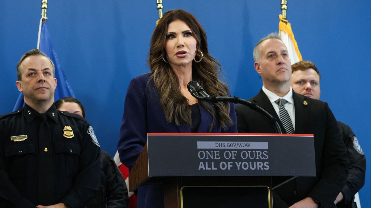A white woman speaks at a podium surrounded by people on either side. The podium says, "ONE OF OURS ALL OF YOURS."