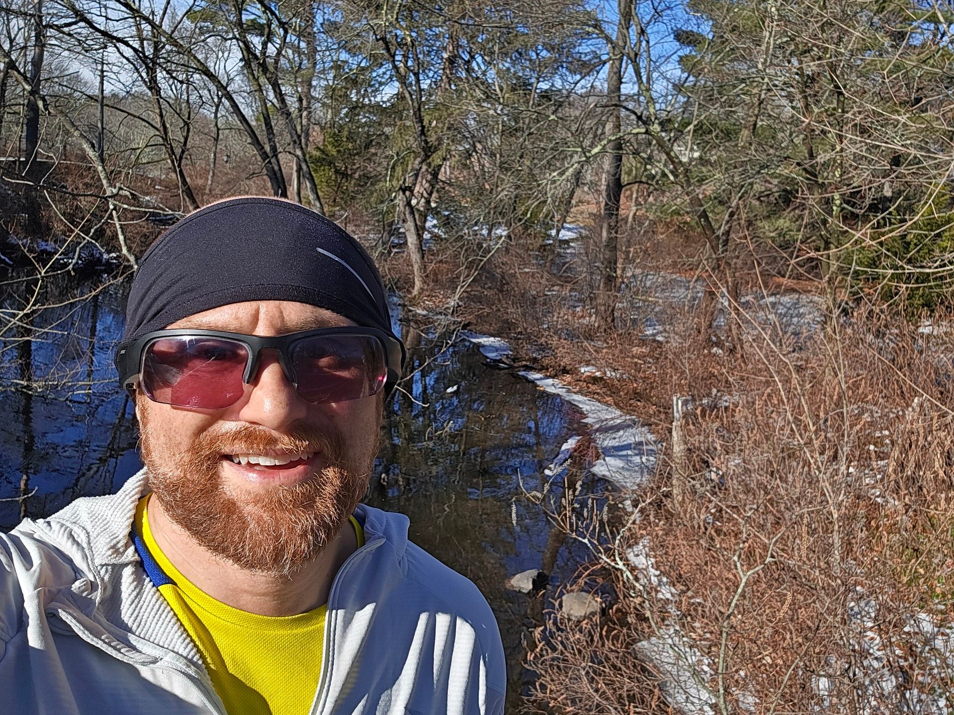 A selfie of me in front of a partially frozen pond, surrounded by bare brambles and trees, on a sunny day. I'm a bald, middle-aged, white man with a red beard flecked with white. I'm wearing a black headband, black sunglasses, and a light aqua partially unzipped running sweater over a yellow and blue running shirt.