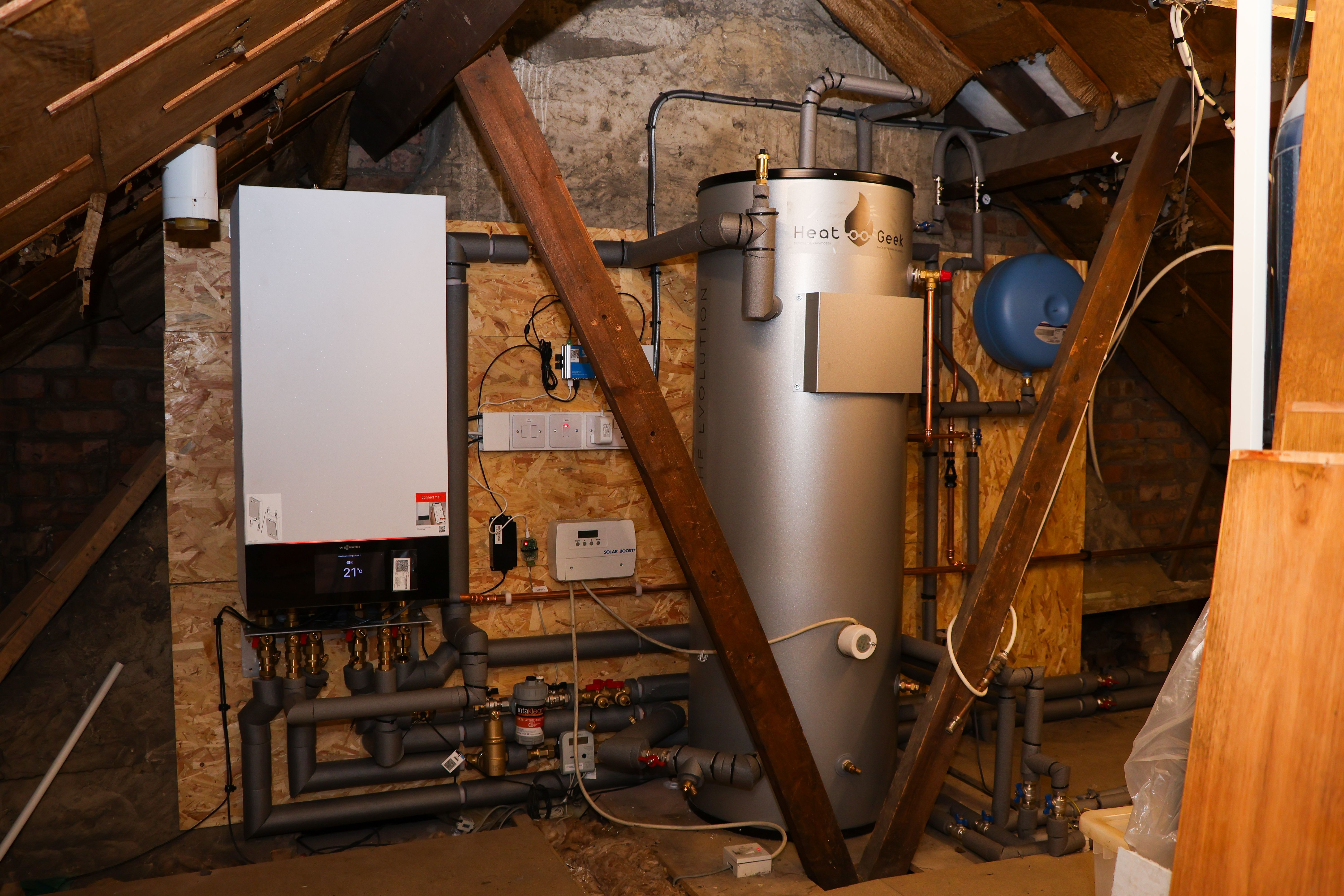 Interior view of a loft-based heating plant room featuring a Viessmann internal controller, a silver Heat Geek Newark 250L hot water cylinder, expansion vessel, and insulated primary pipework.