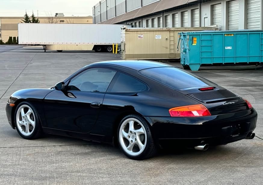 Black 1999 Porsche 996 Carrera 2 with manual transmission, rare "Technology" hollow-spoke wheels, and M030 Sport Suspension, photographed from the rear three-quarter angle in Lakewood, WA. Same owner since 2004, listed for $29,996.