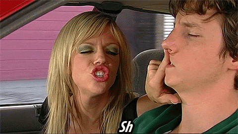 A woman and man in a car and a woman is saying, "shhh" and covering the man's mouth with her hand.
