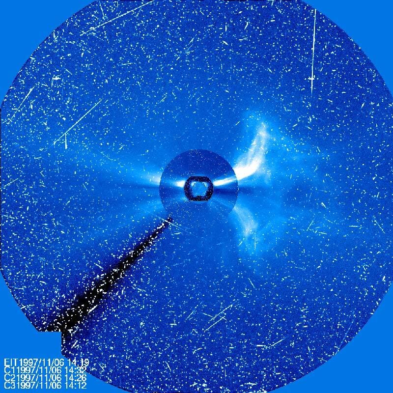 A blue square with a blue circle embedded in it with the Sun at the center, blocked out. Several streamers radiate away from the Sun’s position, and thousands of speckled dot the picture from particle hits.