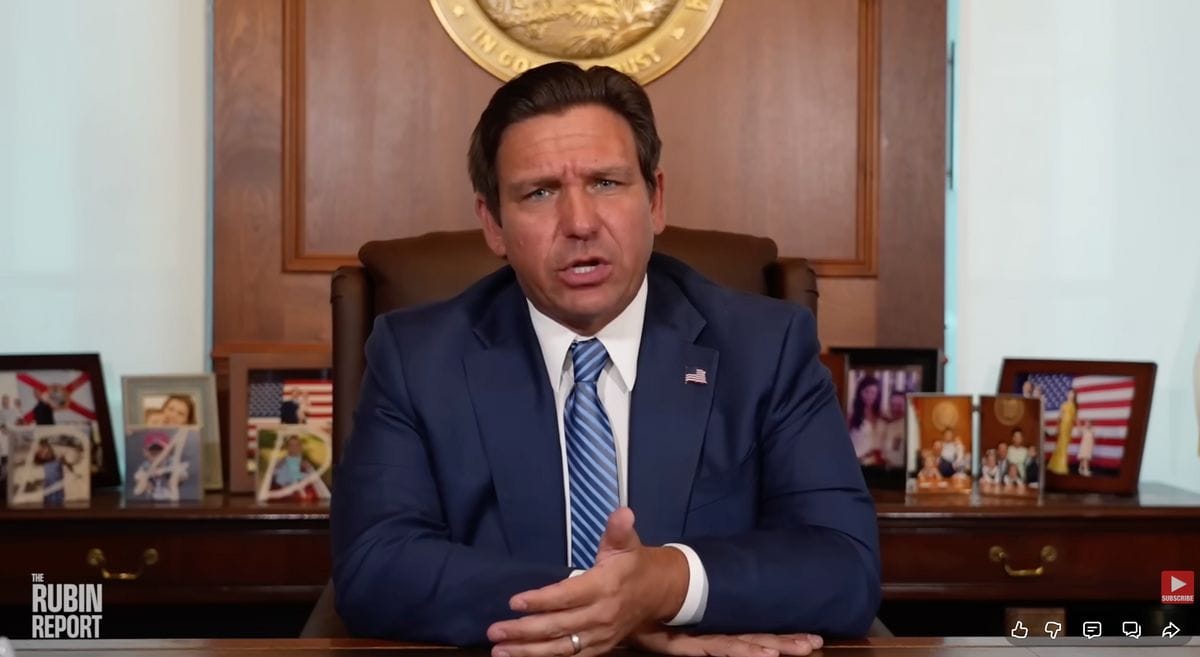 A screengrab shows a white man wearing a blue suit talking while siting at a desk. Various photos can be seen behind him. In the bottom, white text says, "THE RUBI REPORT." A red YouTube subscribe button can be seen in the bottom right corner.