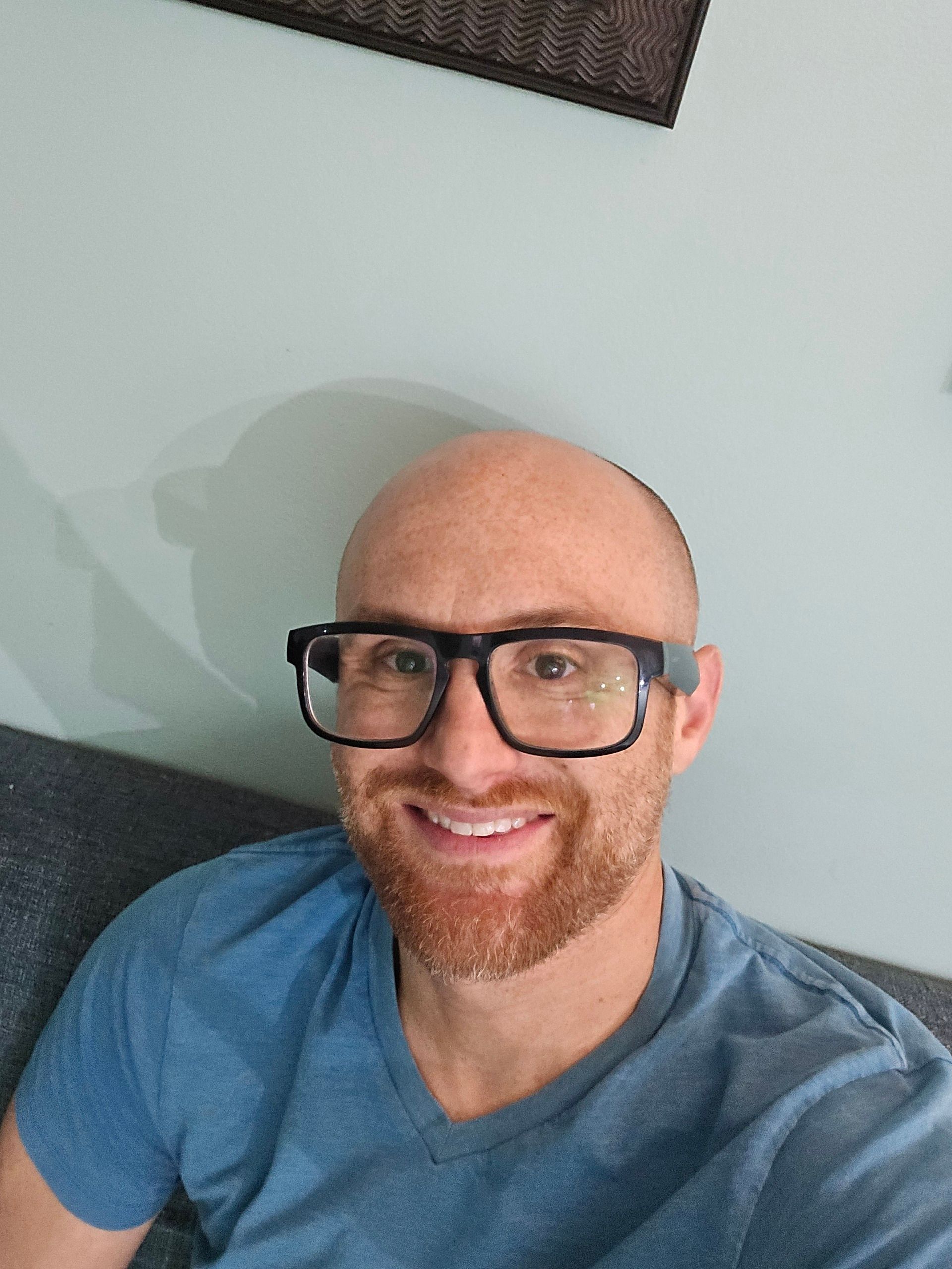A selfie of me sitting on a green couch in front of an aqua wall. I'm a bald, middle-aged, white man with a red beard flecked with white. I'm wearing glasses with thick black rims and a blue t-shirt.