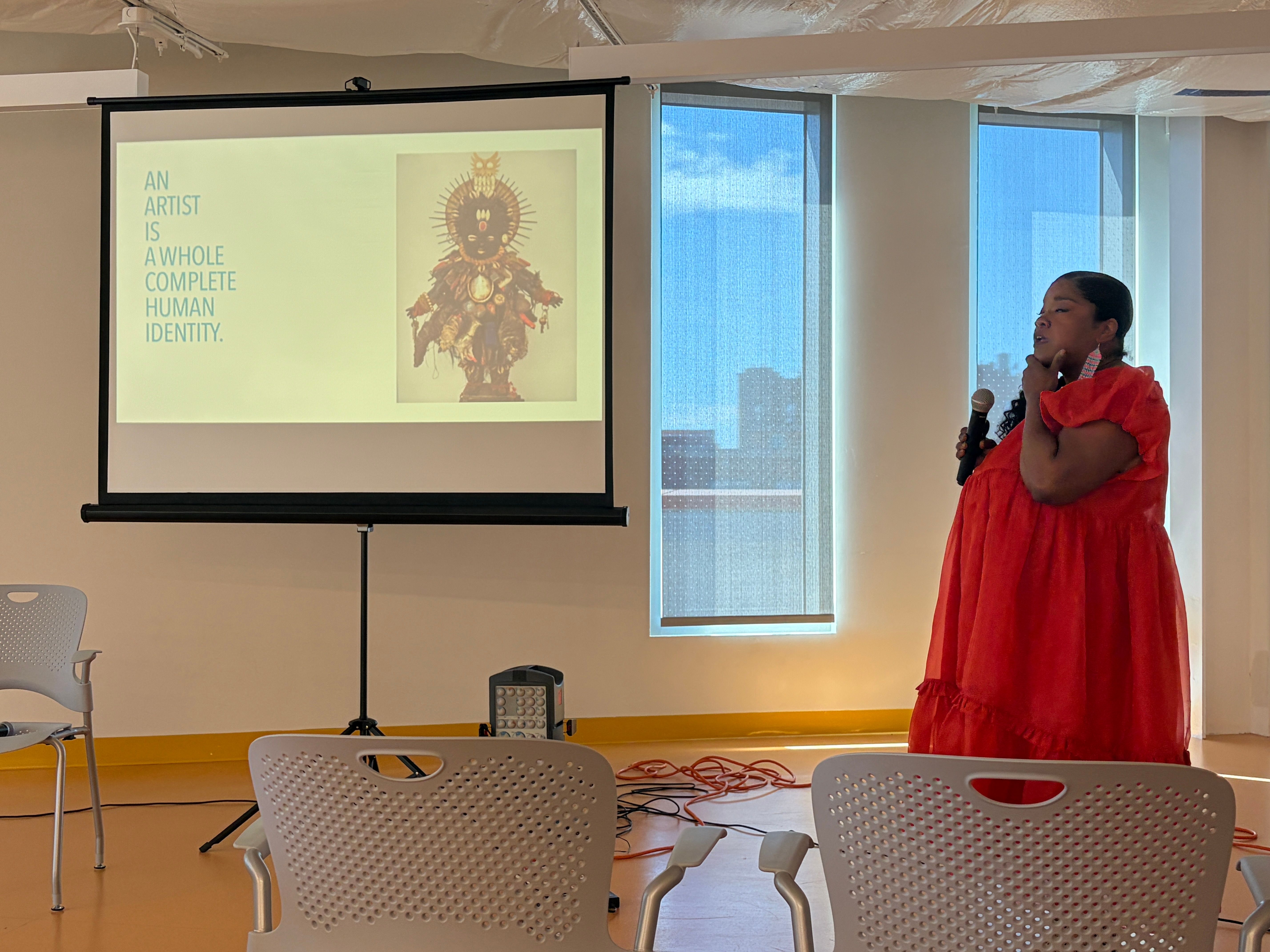 A Black woman in a red dress holds a microphone, presenting beside a screen displaying artwork and text stating "An artist is a whole complete human identity."
