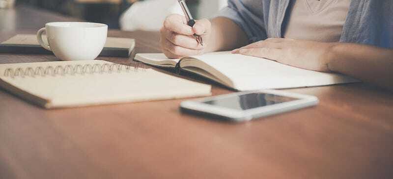 Individual writing notes in a notebook with coffee and phone nearby - illustrating reflection, career planning, and intentional growth.