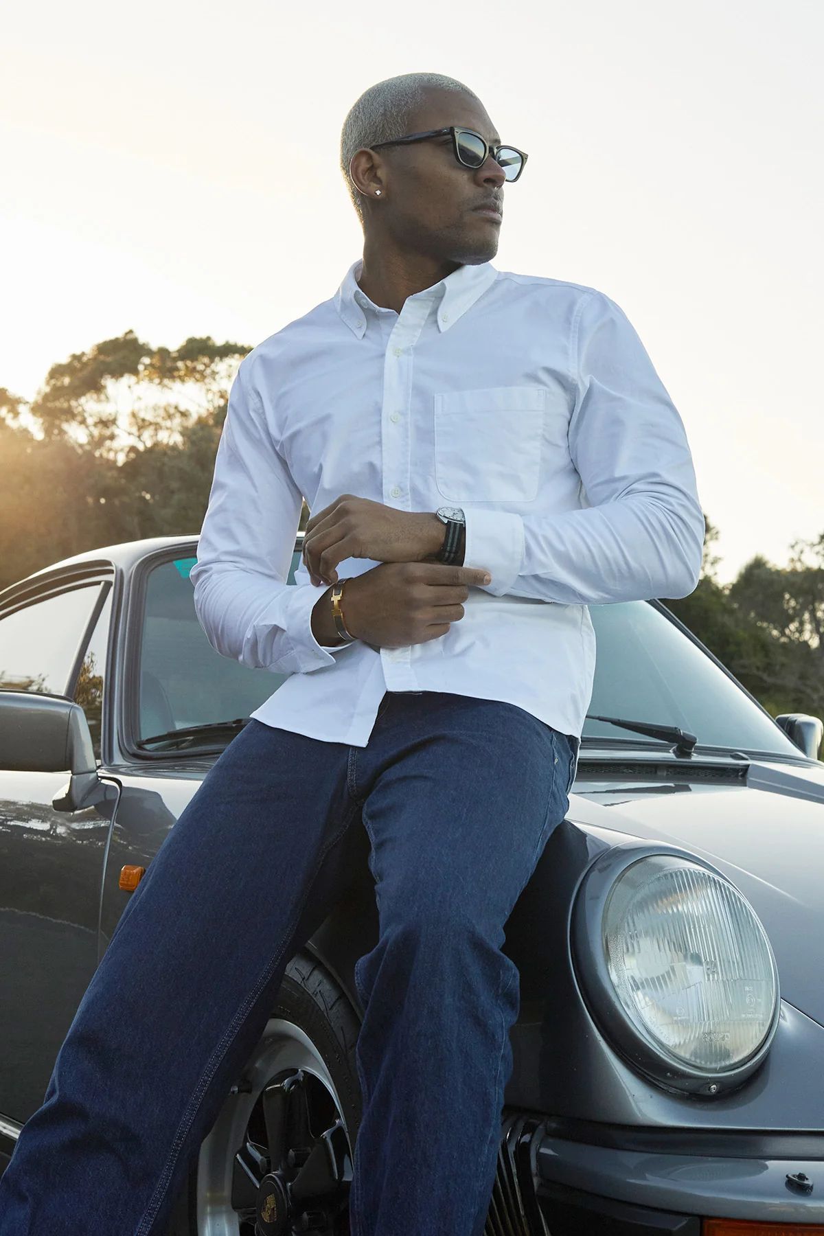 A dark skinned masc presenting person leaning on a car, they are wearing a crisp white custom shirt