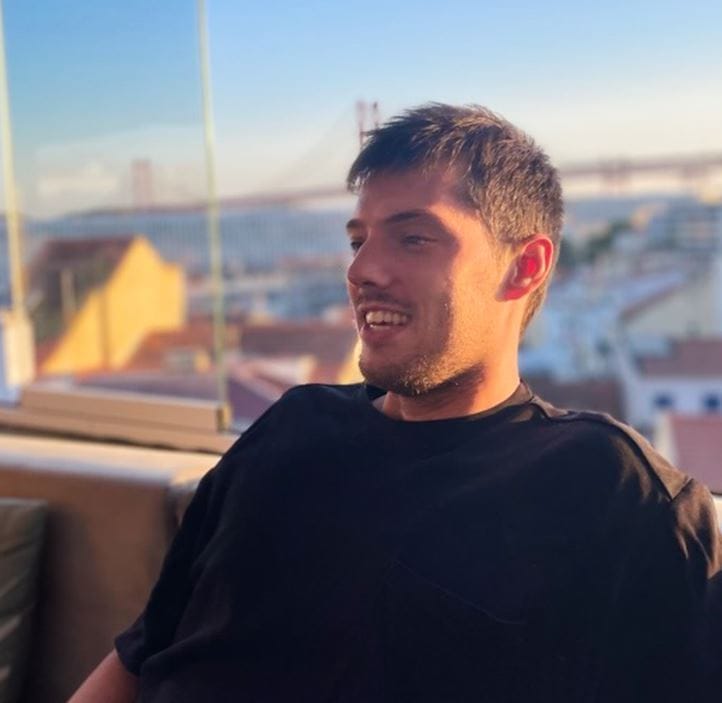 Man in black shirt smiling on balcony with city view, bridge in background, sunny day, modern building. 
