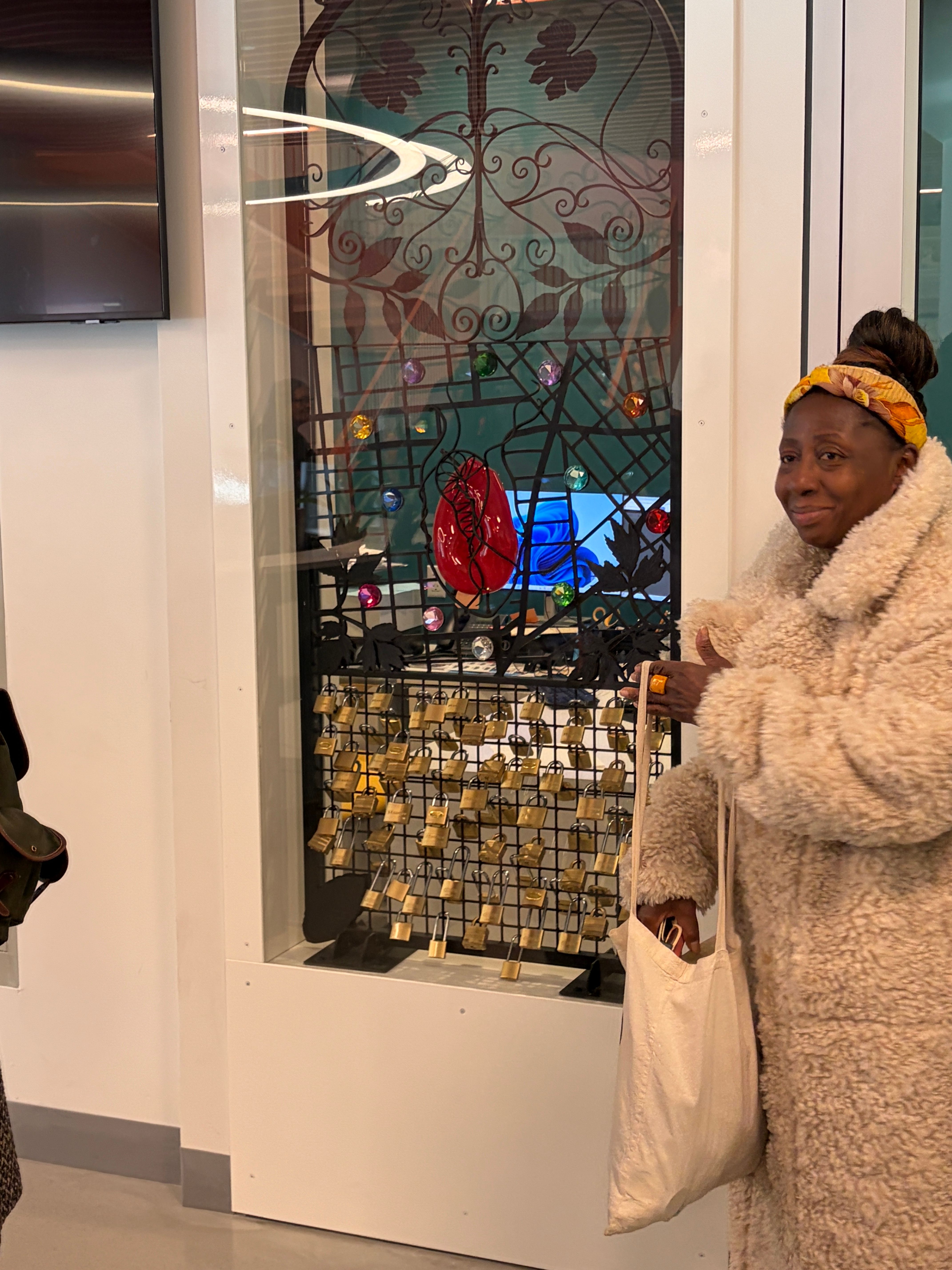A Black woman in a fluffy white coat stands smiling by a decorative iron gate adorned with colorful gems and numerous padlocks. She holds a white tote bag.