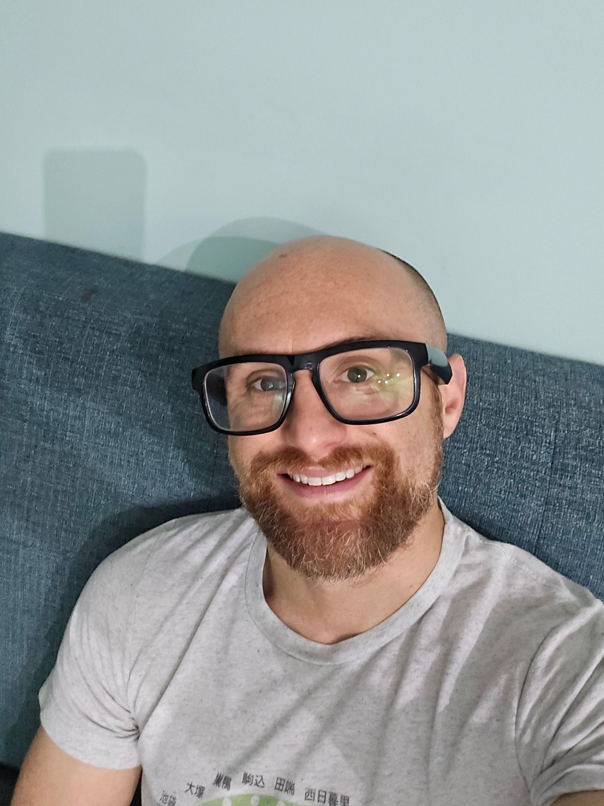 A selfie of me on a green coach in front of a sea foam green wall. I’m a bald, middle-aged, white man with a red beard flecked with white. I’m wearing glasses with thick black rims and a grey t-shirt with a stylized map of the Yamanote Line on it in green.
