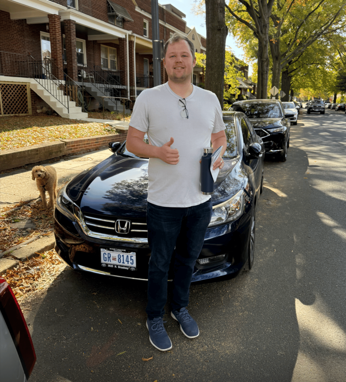 Peter Gustafson standing in front of his dark blue Honda Accord on a tree-lined residential street in Washington, DC, giving a thumbs up before selling the car. A small dog is visible in the background.