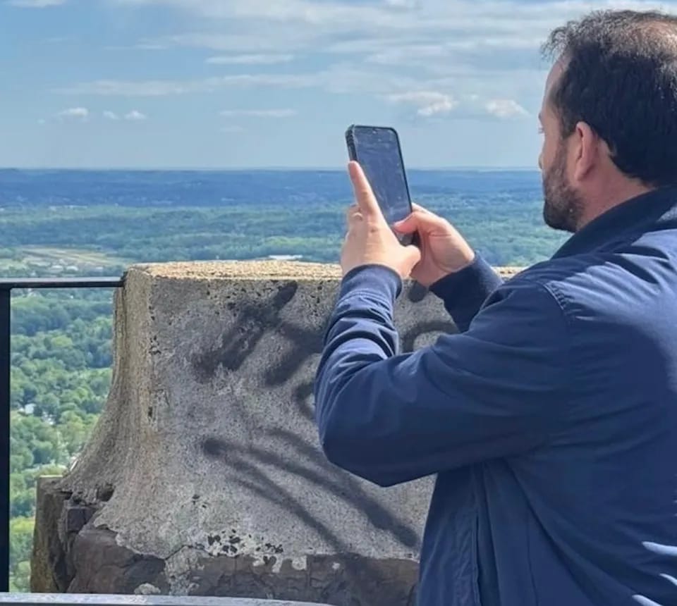 A man in a navy jacket stands at a scenic overlook, holding up his phone to take a photo. Unknowingly, he is blocking the camera with his own hand, preventing the photo from capturing the landscape behind him.