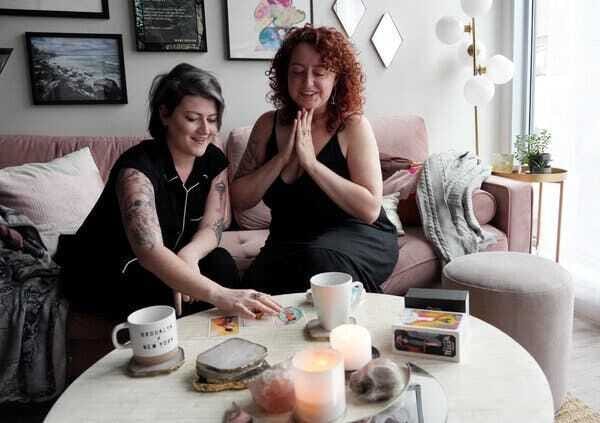 Jeanna Kadlec and her partner Meg Jones Wall, both wearing black, sit in on a dusty rose sofa and read tarot cards. Their living room also has candles, plants and art on the walls.