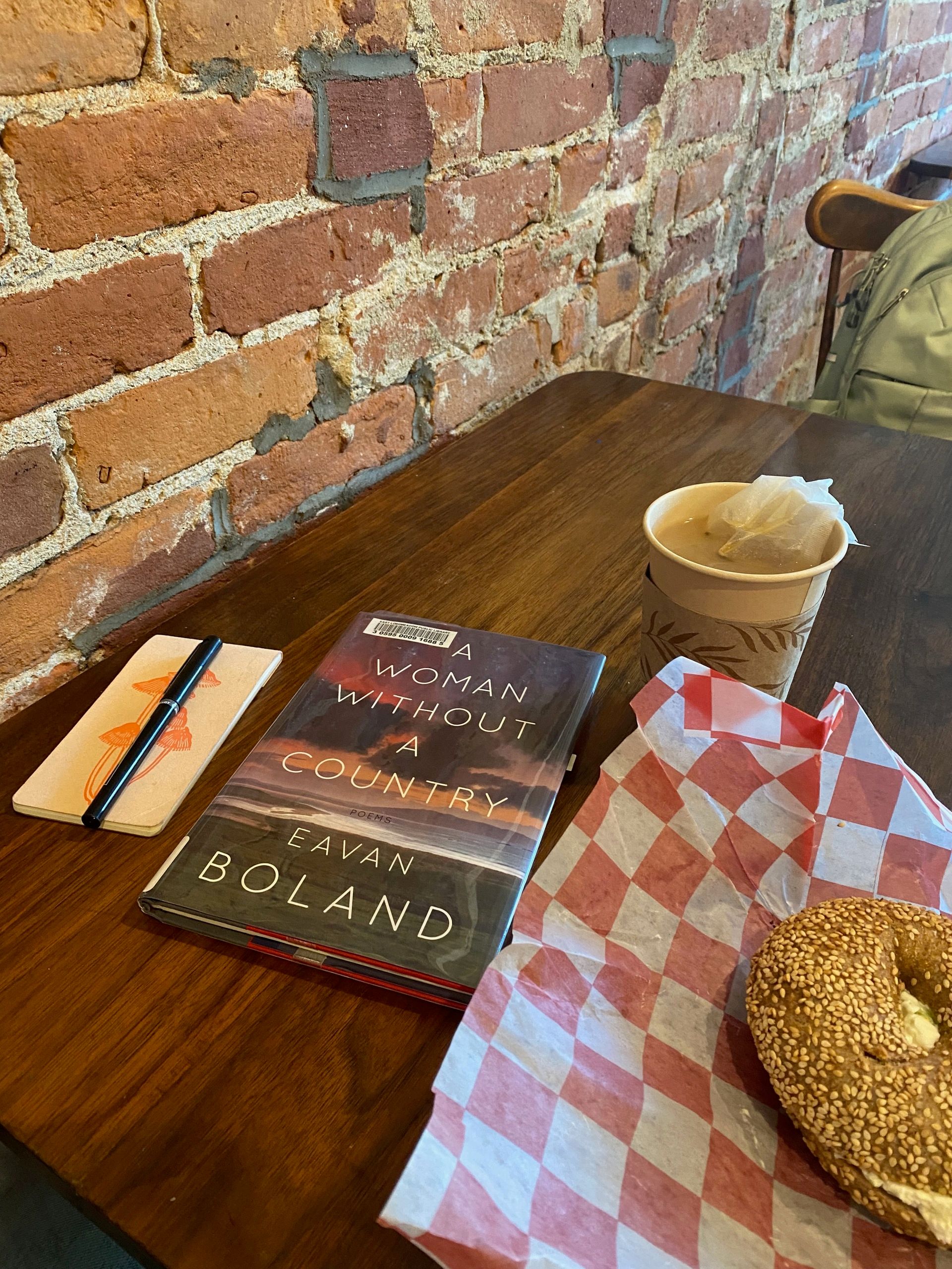 A cafe table with a bagel, a cup of tea, a small notebook with a mushroom on the cover, and a book of poems by Eavan Boland.