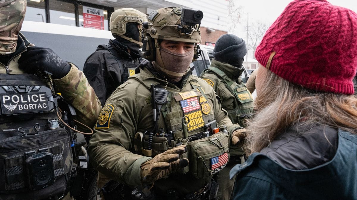 A person wearing a uniform that says BORDER PATROL speaks to someone. They are part of a small crowd of people wearing camoflauge uniforms.