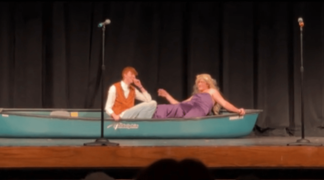 Two teenage boys sit in a boat on a stage. One is dressed as Flynn Ryder and the other as Rapunzel. The former rests his head on his chin while the latter dramatically leans back, blonde wig flowing behind him