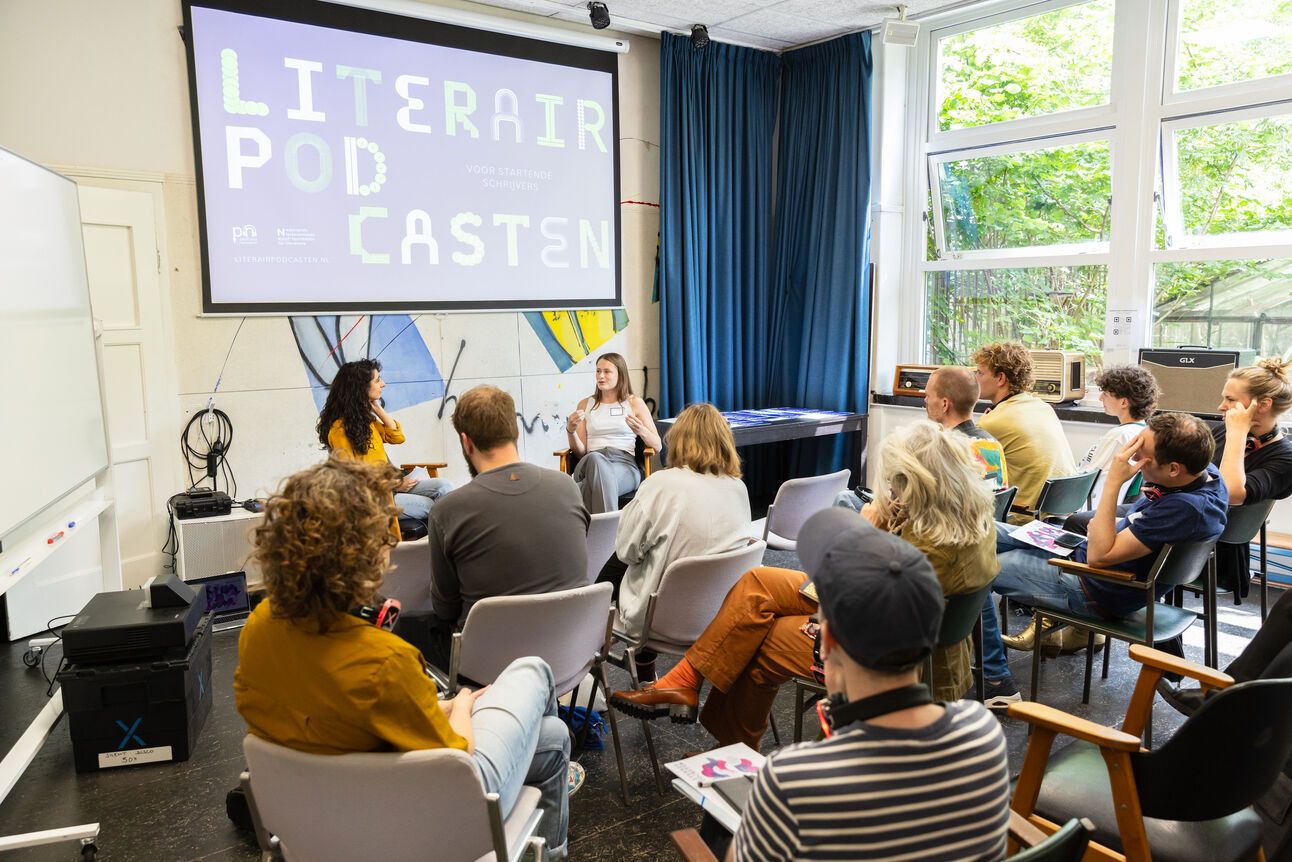 foto van een groep mensen in een klaslokaal, ze zitten op stoelen en luisteren naar twee mensen die voor de groep en een scherm zitten. Op het scherm staat Literair Podcasten en de logo's van het podcastnetwerk en het Letterenfonds. Mina heeft lang zwart krullend haar en een geel shirt en zit links voor de groep, Charlotte de Beus zit rechts en heeft donkerblond halflang haar en draagt een wit topje.