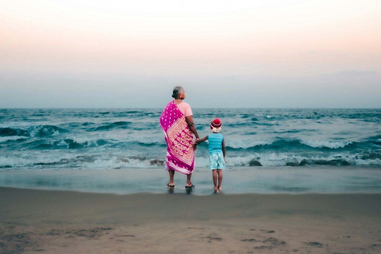 On the shores of a beach, we see the profiles of a grandmother holding the hand of her grandson as the waves lap at their feet